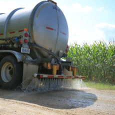 Calcium chloride being spread on an unpaved road next to a cornfield