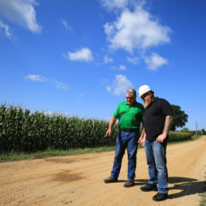 Photo of two man looking at an unpaved road in the summer in front of a corfield